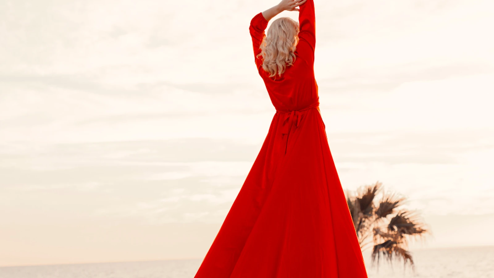 woman with her hands in the air, wearing a floor-length red dress, with her back to the camera, looking out at the ocean.